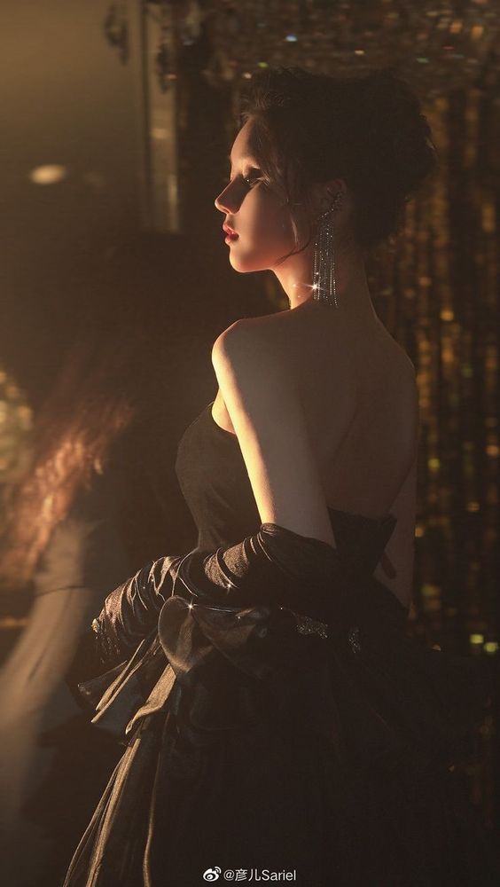 the image showcases a woman elegantly dressed in a black strapless gown and long black gloves, her silhouette illuminated by warm, artificial light. her hair is styled in a sophisticated updo, and she is adorned with long, sparkling earrings. the background suggests an indoor event or party with a shimmering gold curtain behind her, creating a sense of luxury and festivity. the dim lighting casts soft shadows, adding to the dramatic and glamorous ambiance. the perspective is a side view, emphasizing her graceful posture and the flowing lines of her dress.