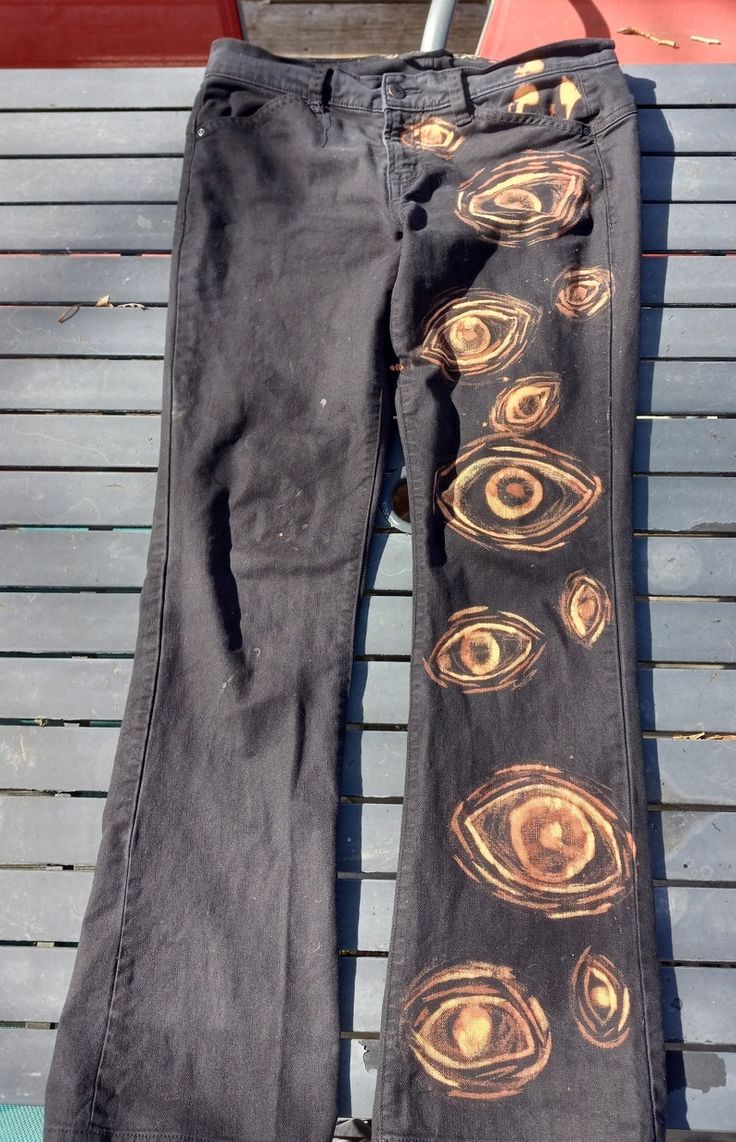 a pair of black jeans with a bleachdye design of multiple yellow eyes on the right leg. the jeans are laid out on a wooden deck and the right leg is pulled up to reveal the design more clearly. the jeans are a flared style.
