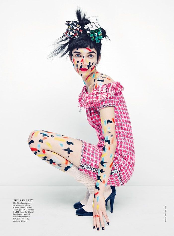a female model is dressed in a pink and white dress with a colorful paint design, black heels and black and white painted makeup. she is crouched down with one hand on the floor and the other holding her body up. she is wearing a black and white hair piece, with a colorful paint design as well. the photo appears to have been taken in a studio on a white backdrop. the image is an editorial photoshoot of a model.