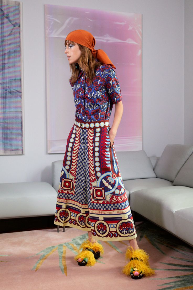 a full shot of a fashion model wearing a vibrant, patterned outfit. the outfit consists of a shortsleeved, blue and floral patterned shirt tucked into a highwaisted, wideleg palazzo pant. the pants feature a complex, multicolored geometric and floral design.  the model also wears a burnt orange headscarf and bright yellow fuzzy slippers. she is standing in a modern living room with a light grey sofa and a pink area rug. the backdrop includes modern wall art. the overall style is bohemian and retro.