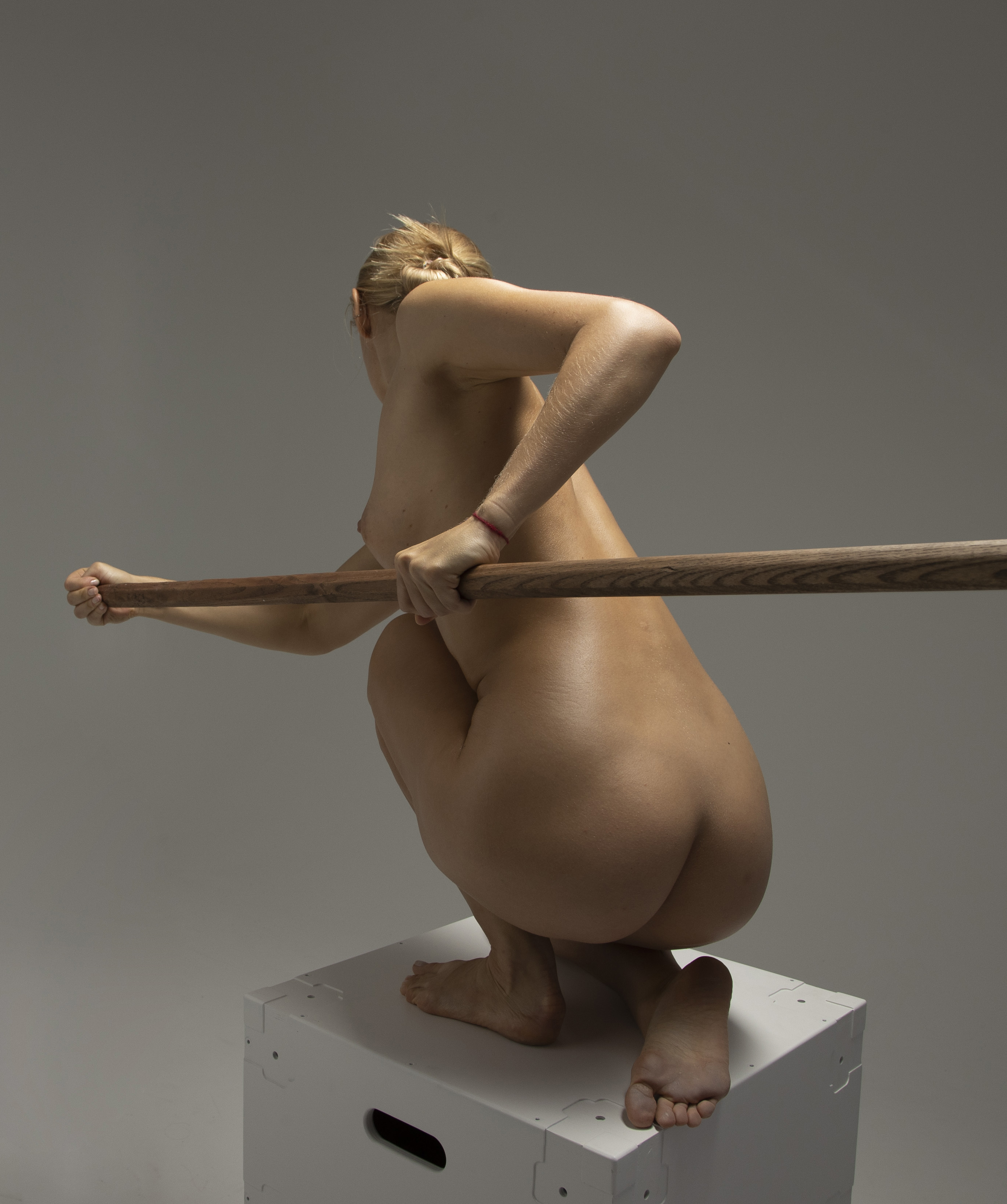 a nude woman is squatting on a white box, holding a wooden bar horizontally in front of her. the photo is taken from behind, showing her back and the back of her legs. her blonde hair is tied up, and she has her arms raised slightly to grip the bar. the background is a plain grey studio backdrop. the lighting is soft, highlighting the contours of her body.