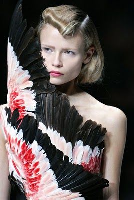 a woman with long blonde hair is modeling a black, white, and red feathered dress.  she is looking at the camera with a slightly sad expression.  the feathers are arranged in a winglike pattern. the image has a dark background and a simple composition. the woman is wearing minimal makeup, and she is styled with a simple hairstyle.
