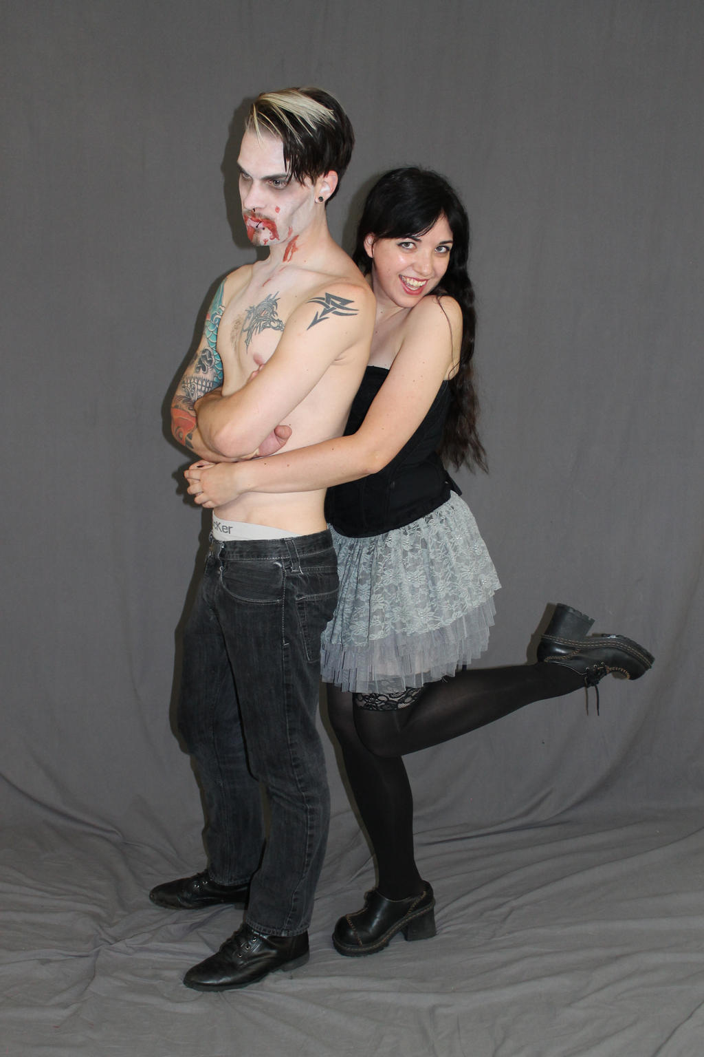 a full shot of a couple posing against a gray backdrop. the man, with zombielike makeup and tattoos, is shirtless, wearing dark jeans, and has light and dark hair.  the woman, with dark hair, is wearing a grey layered skirt, black tights, and black boots. she is embracing the man around his torso. the image has a dark, alternative style.