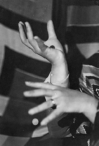 a closeup black and white photograph captures the expressive hands of a woman. her hands are elegantly posed, one palm up and the other overlapping, suggesting a gesture of communication or artistic expression. she wears a ring on the lower hand. the background is blurred but shows a patterned fabric, perhaps a garment or textile. the image is high in contrast, with deep shadows and bright highlights, giving it a dramatic and artistic feel. the style is reminiscent of vintage or retro photography, possibly from the mid20th century.