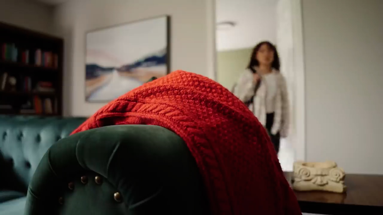 a woman is walking away from the camera, out of a living room. the focus is on the foreground, with a soft red knit blanket draped over a dark green velvet armchair. the armchair is partially visible, showcasing its button detailing. to the left, a sliver of a matching couch is seen.  behind the chair, a doorway leads to a brighter hallway where the woman in a white shirt is exiting.  the living room is decorated with a large painting on the wall and a bookshelf filled with books is visible in the background. the room is bathed in soft, natural light.