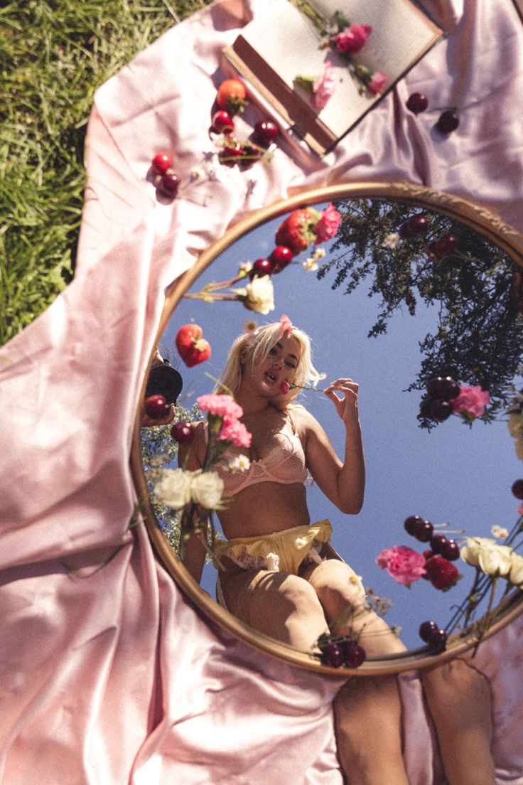 the image depicts a blonde woman in pink lingerie reflected in a large, round, goldframed mirror.  the mirror is placed on a pink silk fabric outdoors in a grassy area, surrounded by scattered cherries and a floral arrangement. the reflection shows the woman upside down, against a bright blue sky; the overall feel is romantic and pastel. the scene is suggestive of a summer picnic or outdoor boudoir setting. the flowers and cherries add to the feminine and summery ambiance.