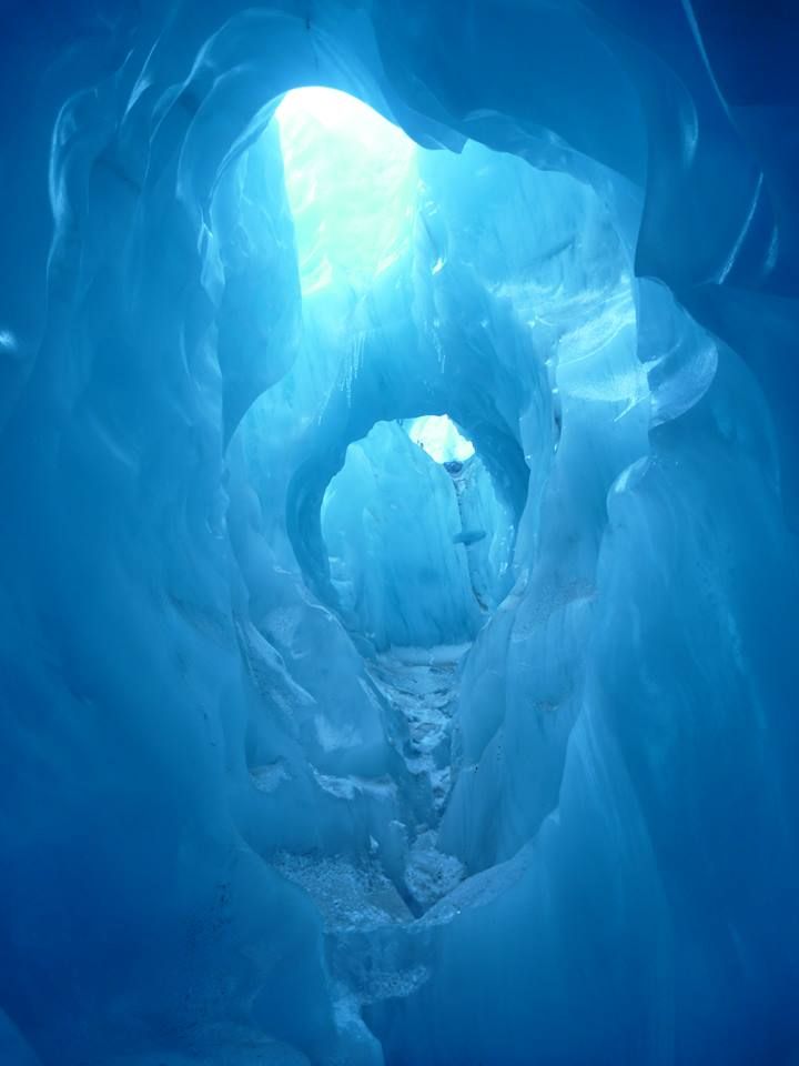 an image of a natural ice cave. the cave is made of blue ice and has a tunnellike shape. light shines through openings at the ends of the tunnel, creating a bright and ethereal atmosphere. the walls and ceiling of the cave are irregular and textured, with some icicles hanging down.