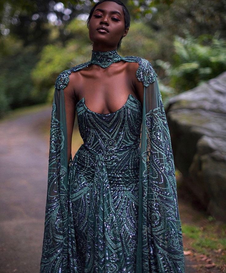 the image showcases a darkskinned model wearing a stunning, floorlength dark green gown. the dress features intricate sequined embroidery and beading throughout, creating a luxurious and sophisticated look.  the gown has a halter neckline with a highneck cape that flows elegantly from the shoulders. the cape sleeves are sheer and add a touch of ethereal beauty. the model is posing outdoors in a forest setting, which provides a dramatic and moody backdrop that complements the dress's color and design.  the overall style is elegant and regal.