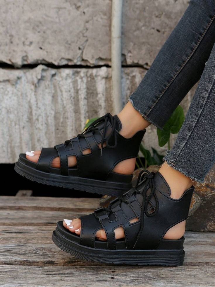 a closeup shot shows a person wearing black laceup gladiatorstyle platform sandals. the sandals have an opentoe design and the person is wearing dark denim jeans with a frayed hem. the background includes weathered concrete and wooden planks.