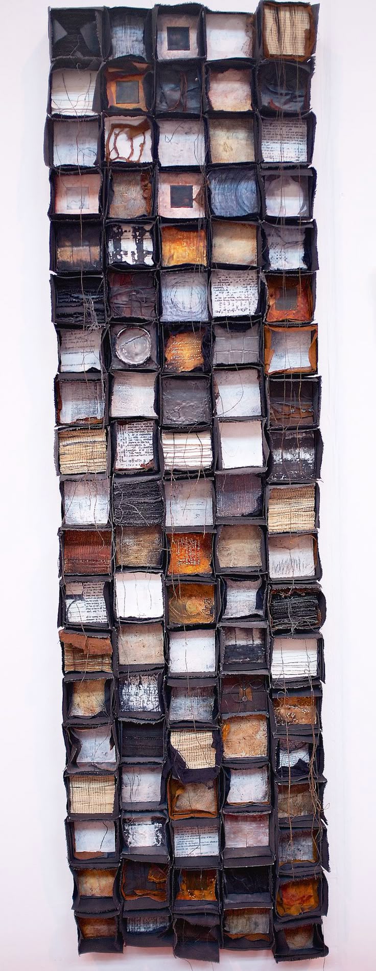 the image shows a tall, rectangular mixed media collage artwork made of small, darkframed boxes. each box contains different materials such as paper, fabric, bits of books, and metal. the overall impression is of a textured, gridlike arrangement with a monochrome or muted color palette, primarily in shades of brown, black, and white. the piece evokes a sense of recycling or repurposing, possibly with an abstract or conceptual theme. the materials within each box vary in texture and pattern.