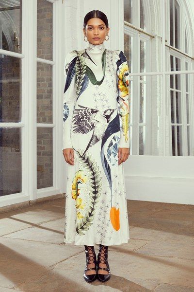 a full shot of a model wearing a white midi dress with a vibrant floral and bird print. the dress features a high neck and long sleeves, with a fitted silhouette that flares slightly at the hem. the model is posed against a backdrop of large windows in a bright, airy space. she is wearing black laceup heels, completing the sophisticated and elegant look. the overall aesthetic of the image is clean, modern, and highfashion.