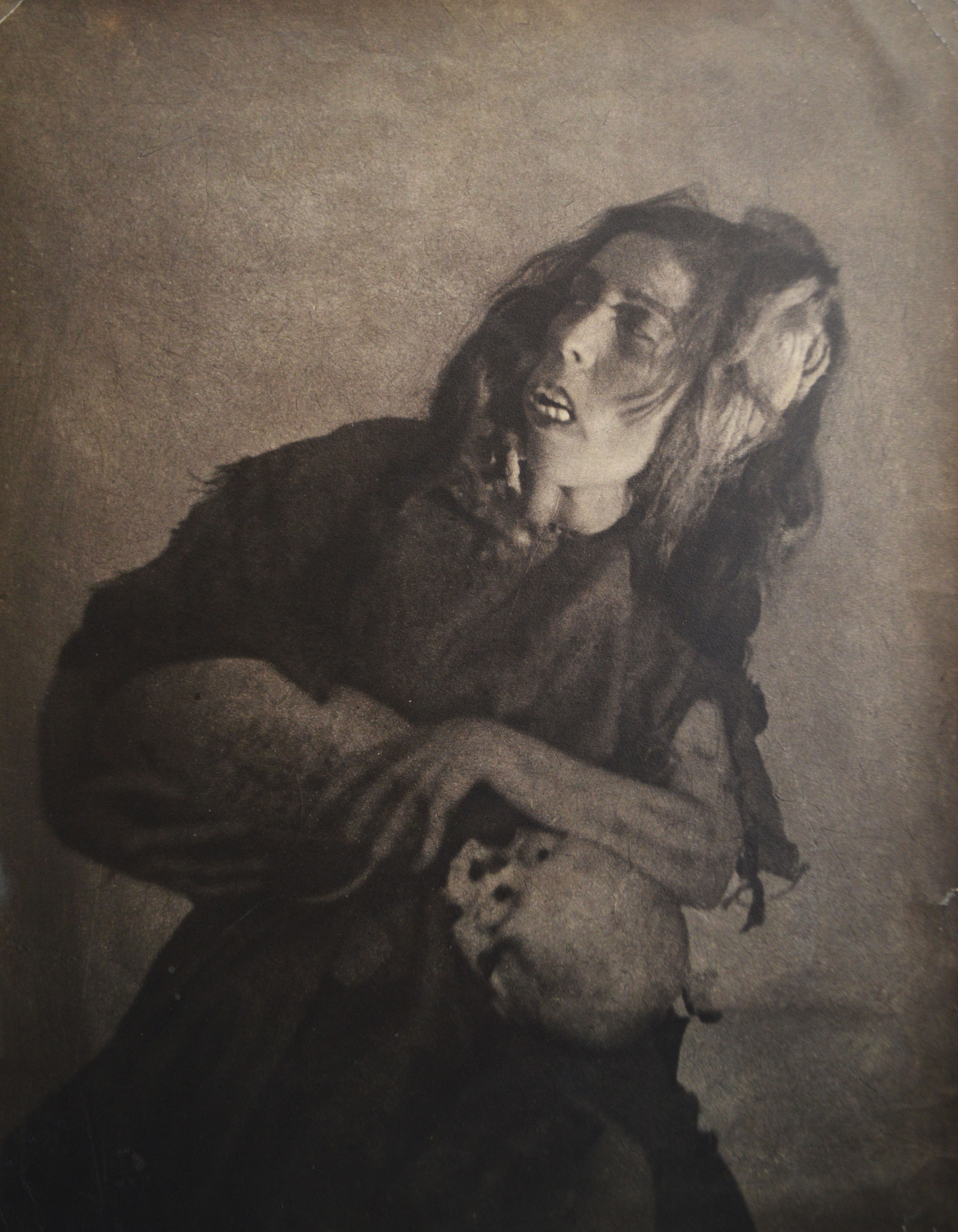 sepiatoned, vintage photograph depicts a woman in a dark, possibly theatrical, costume. her face is partially obscured by shadows, yet her expression appears one of intense pain or anguish.  her hands are clasped tightly in front of her, and her posture suggests a state of deep distress or despair. the overall style of the image is dramatic and moody, with the dark tones and intense expression creating a haunting atmosphere.  the image's quality suggests it's an antique photograph, possibly from a play or theatrical production.