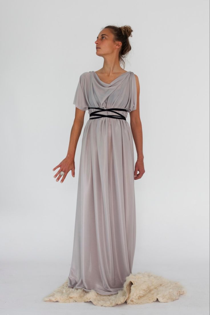 a fulllength studio shot of a woman wearing a long, flowing grey grecianstyle dress. the dress has a cowl neck and a black belt.  she is standing on a light creamcolored fluffy rug, against a plain white background. the model's pose is simple and elegant. the overall aesthetic is minimal and sophisticated.