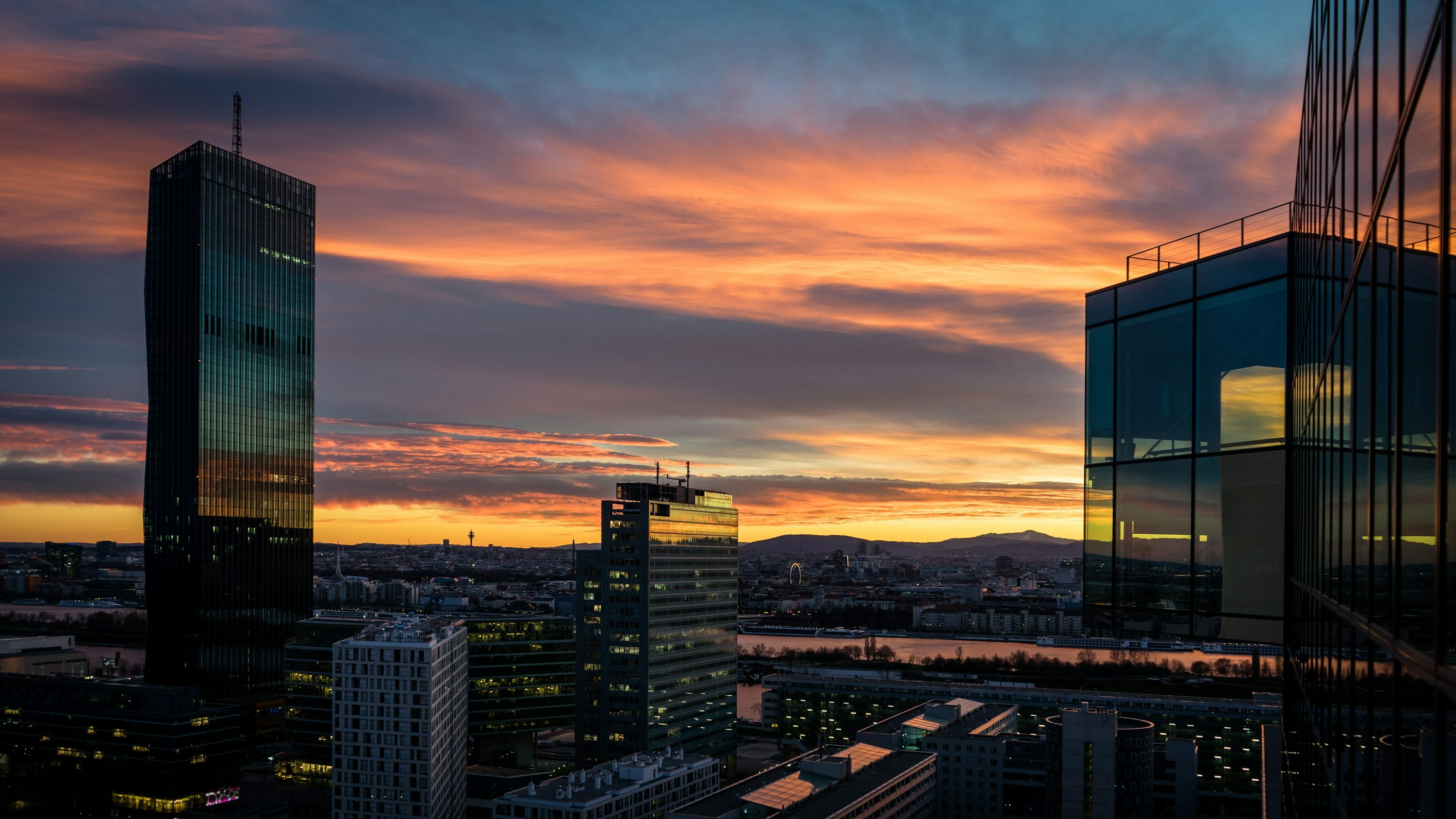 the image depicts a cityscape at sunset, with several skyscrapers standing prominently against a vibrant orange and purple sky filled with clouds. the buildings reflect the colors of the sky, adding depth and dimension to the urban scene. the overall impression is one of modern architecture meeting natural beauty.