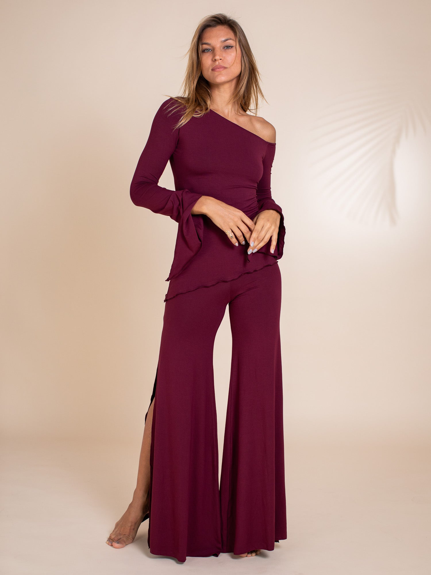 the image shows a fulllength shot of a woman model elegantly posing in a marooncolored outfit. the outfit consists of a longsleeve, offshoulder top with ruffle sleeves and wideleg pants featuring side slits. the pants flare out dramatically from the knee, creating a bellbottom silhouette. the fabric appears to be a soft, flowing material. the model's hair is styled in loose waves that fall past her shoulders. the background is a light beige, ensuring the outfit is the focal point. the overall aesthetic is elegant and stylish.