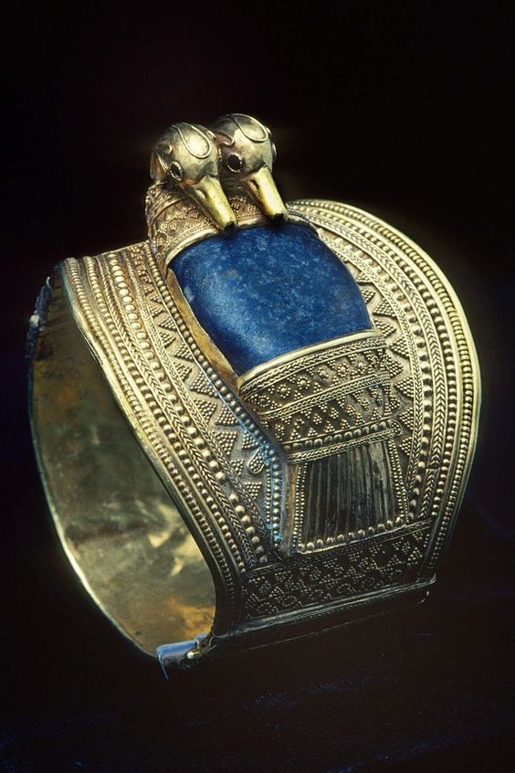the image showcases a gold bracelet adorned with lapis lazuli and featuring two bird heads. the bracelet is intricately crafted with patterns and textures. the dark background emphasizes the bracelet's details, and the studio lighting highlights the richness of its colors and the craftsmanship involved in its creation.