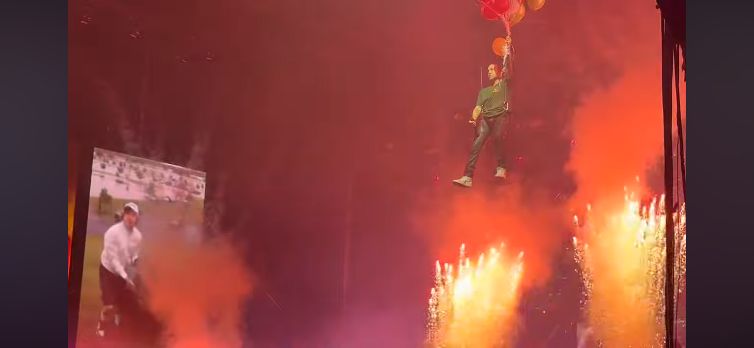 a performer is suspended in midair, being hoisted up above a crowd during a concert. the performer is dressed in dark clothing and is holding onto a harness attached to balloons. in the background are multiple fireworks displays, and redcolored smoke.