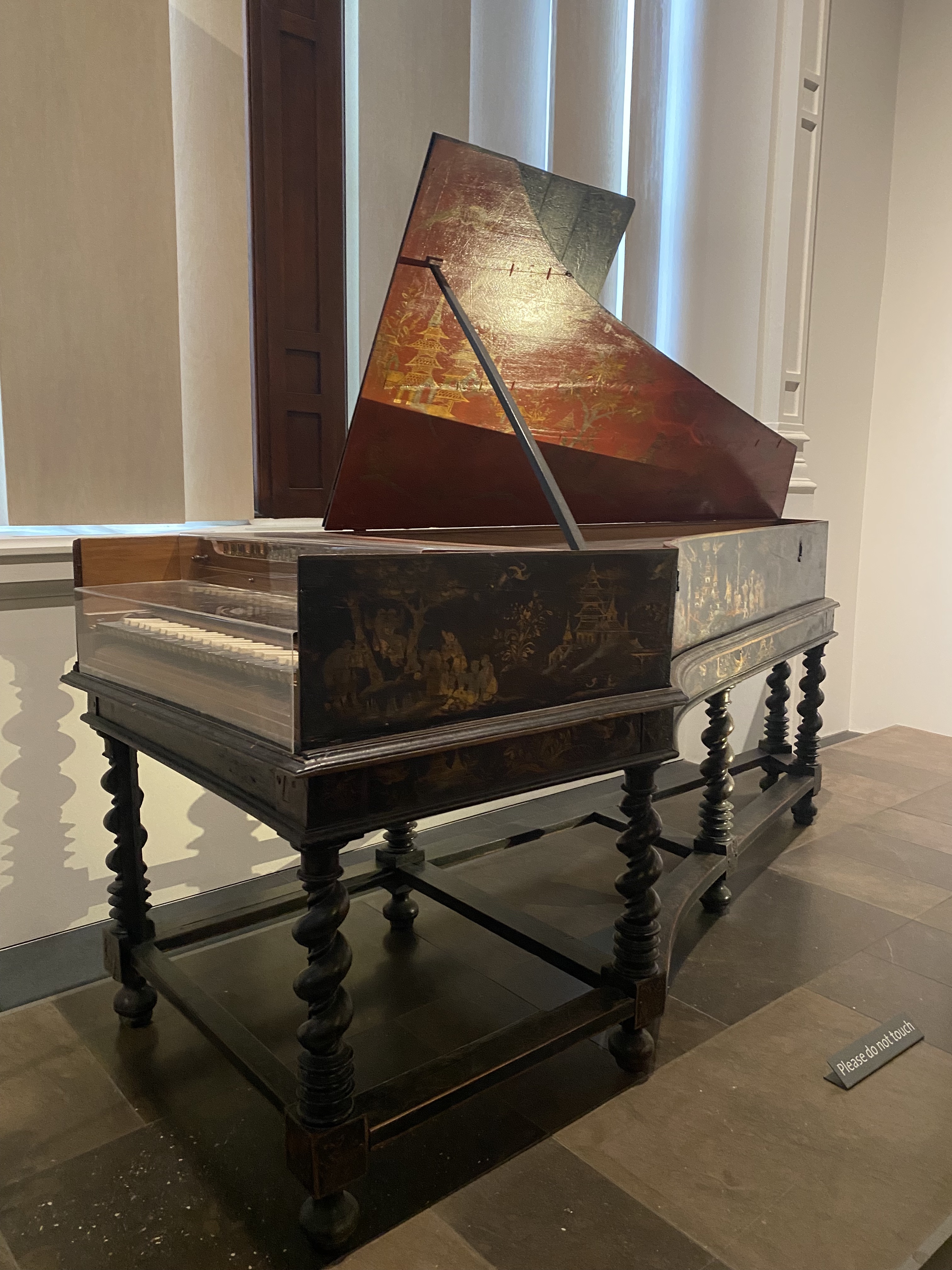 a highangle shot of an antique harpsichord displayed in a museum setting. the instrument has a dark, ornate stand with twisted legs and a lower shelf. the harpsichord's body is painted with intricate designs, and the soundboard has a red and gold coloration. a transparent display case covers the keyboard. the background features a window with diffused light and a sign that reads "please do not touch."