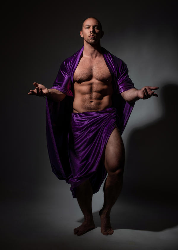 a muscular, bald man is posing in a studio setting against a dark background. he is draped in a rich, purple cloth that resembles a toga or a shawl. his muscular physique is clearly visible, and his expression is serious and confident.  the lighting is dramatic, creating shadows and highlighting his features and the texture of the cloth. the overall style evokes classical sculpture or a depiction of a mythological figure.