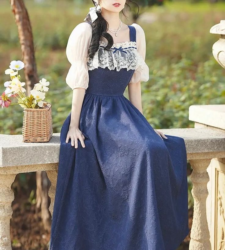 a woman wearing a vintageinspired blue dress is sitting on a stone balustrade in a garden setting. the dress features a square neckline, delicate lace detailing on the bodice, and a flowing skirt. the soft, natural lighting and surrounding greenery create a romantic and feminine atmosphere.