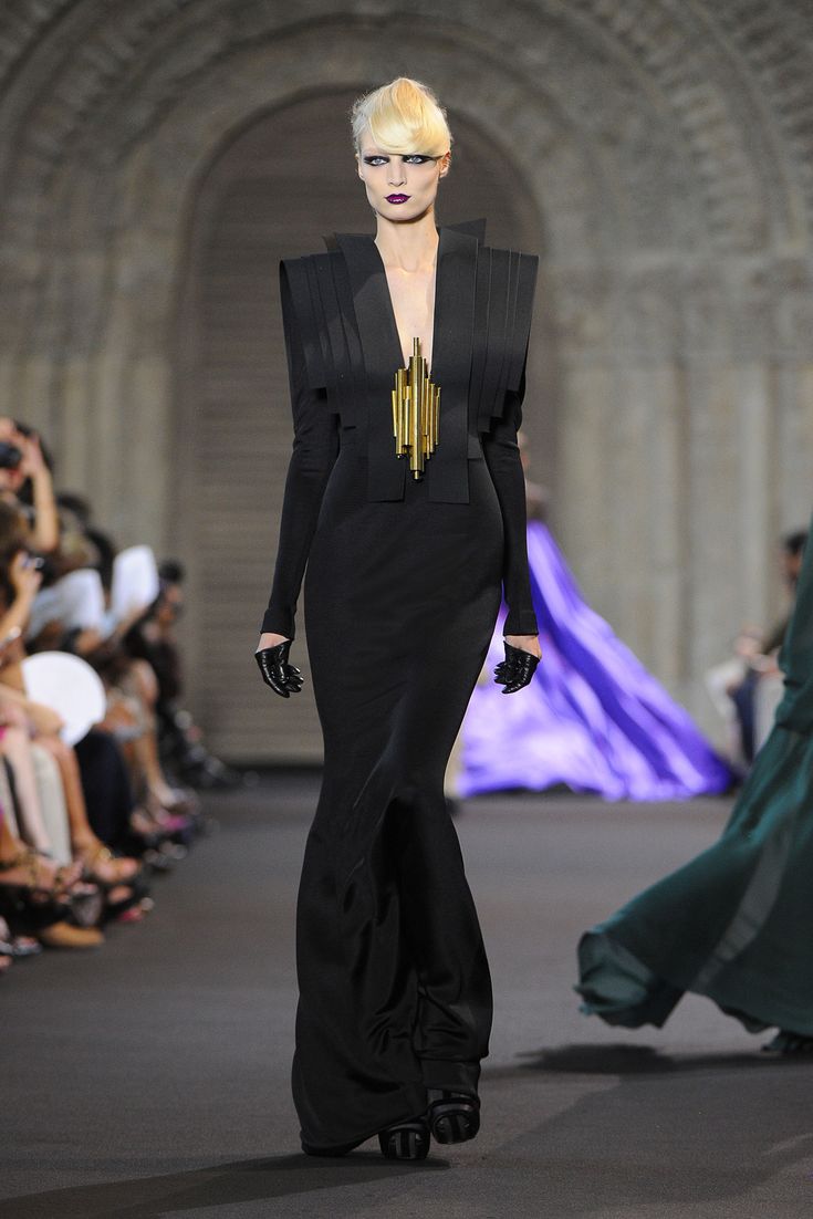 a model with blonde hair and dark makeup walks down a runway wearing a long, black, elegant gown. the dress has structured shoulders and a gold geometric necklace. the model wears black gloves and high heels. the background shows an audience and other models in different colored gowns. the setting appears to be a fashion show.