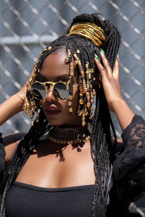 a young african woman with long braids adorned with gold beads, wearing a gold choker, and sunglasses, stands in front of a chain link fence. her hair is styled in a high ponytail with multiple gold bands.  she is wearing a black offtheshoulder top and a black lace jacket. her hair is styled in a high ponytail with multiple gold bands. the woman is looking at the camera with a confident expression, and she is wearing a black choker necklace and a pair of round, goldrimmed sunglasses.