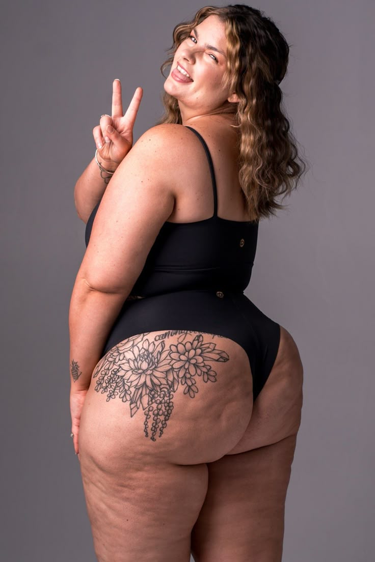 a plussize woman wearing black lingerie stands against a gray background, flashing a peace sign and sticking her tongue out in a playful expression. she has curly hair pulled back behind her ear and a large floral tattoo on her thigh. the image promotes body positivity and selfacceptance.