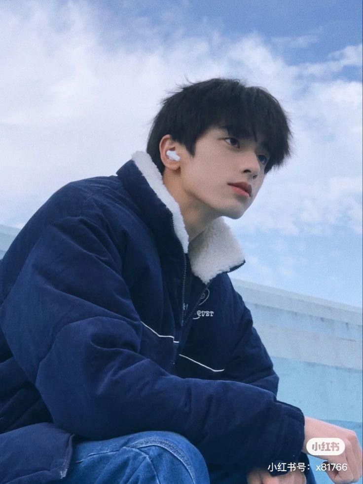 a young asian man wearing a blue puffer jacket sits outdoors with a contemplative expression. the background features a bright blue sky with fluffy white clouds. the image is taken from an eyelevel perspective, capturing the man's upper body and a portion of his legs. he wears white earbuds in his ears and has a relaxed posture. the overall feel of the image is youthful and serene.