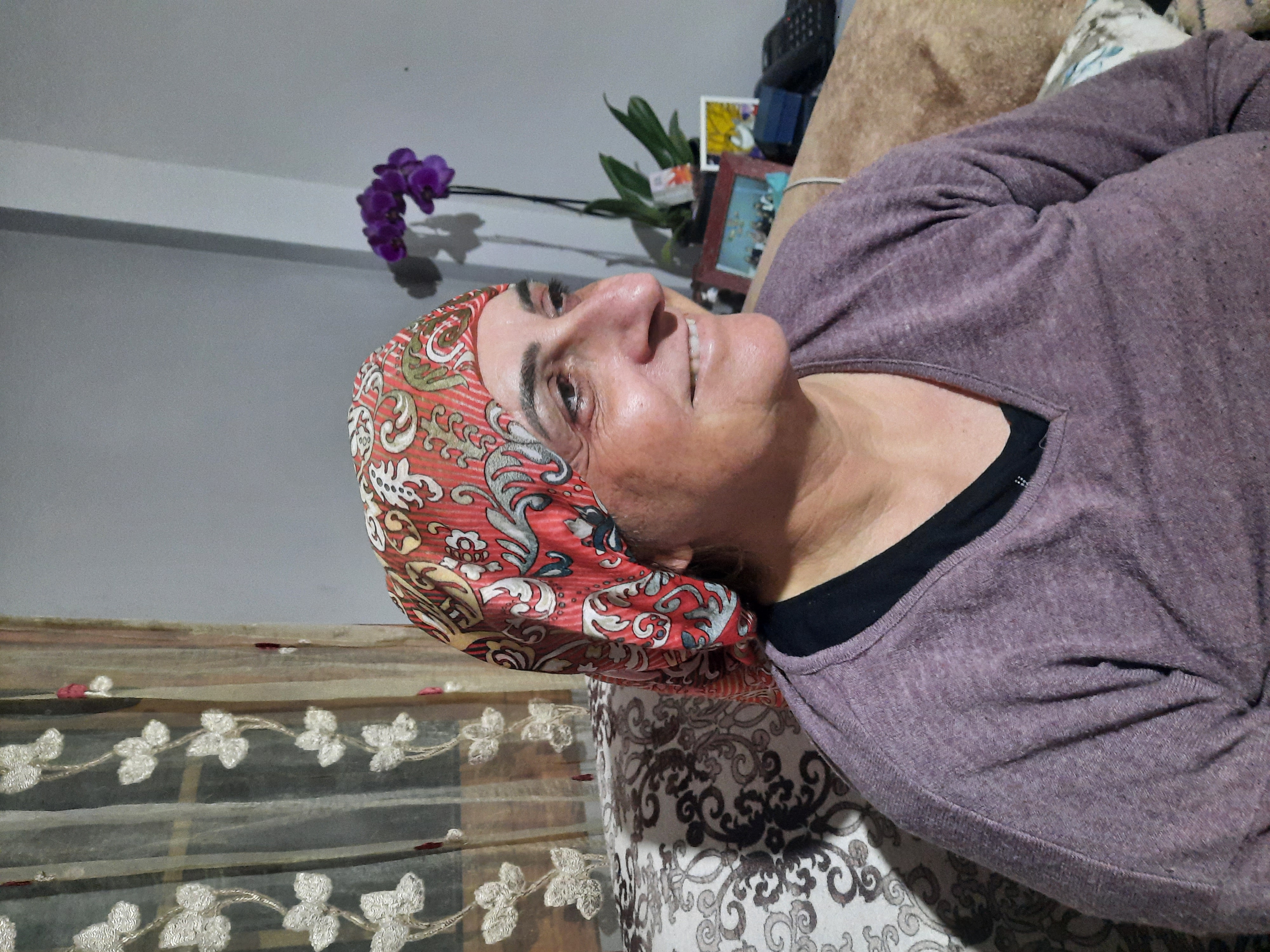 a woman wearing a floral patterned headscarf and a purple sweater sits on a couch indoors. an orchid is visible in the background, along with decorative elements such as curtains and a floral patterned couch. she is smiling and looking to the side.