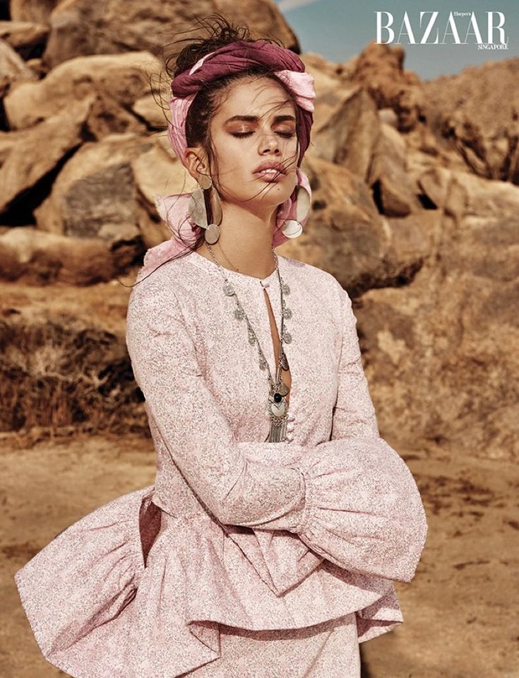 the image is a fashion editorial shot from harper's bazaar singapore. it features a female model wearing a light pink floral peasantstyle dress with bell sleeves and a ruffled hem. she has on a  pink and maroon head wrap styled as a turban, large silver earrings, and a layered silver necklace.  her eyes are closed, and she appears serene and relaxed. the background is a sundrenched desert landscape with large rocks, creating a bohemian and romantic atmosphere. the overall aesthetic is high fashion, with a focus on the clothing and the model's beauty.