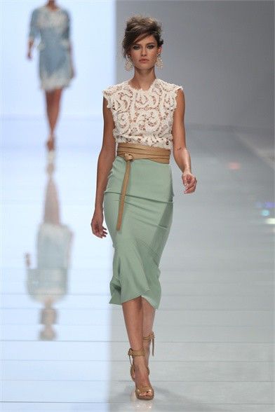 a model walks down a runway wearing a stylish outfit. the outfit consists of a white lace sleeveless top paired with a light sage green pencil skirt.  a light brown leather belt cinches the waist. the model also wears tan heeled sandals and has her hair styled in a chic updo.  another woman in a light blue dress is visible in the background, slightly out of focus. the overall setting suggests a fashion show.