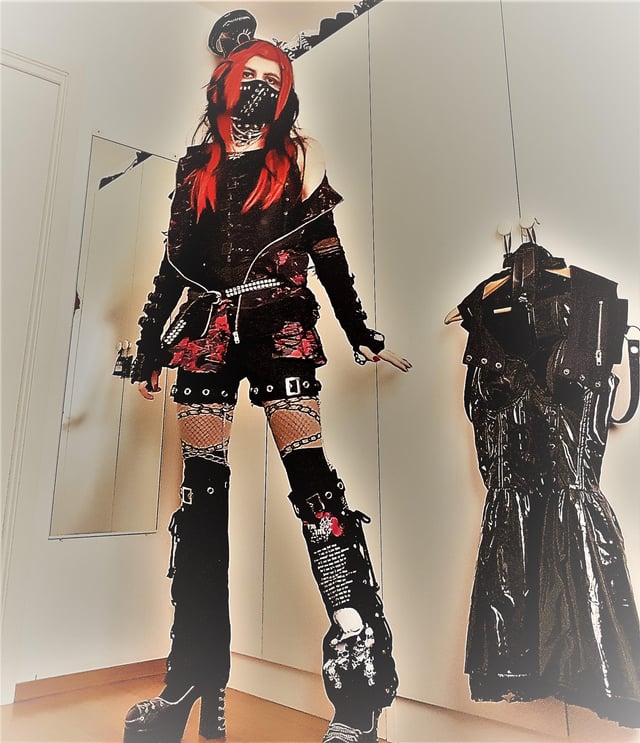 a person is taking a mirror selfie of their outfit. they are wearing a black and red visual kei inspired outfit, consisting of a black jacket over a red and black dress, with black shorts over fishnet tights, and black platform boots with buckles and straps. they have red hair and are wearing a face mask. the background is a bedroom with white walls and a white closet.