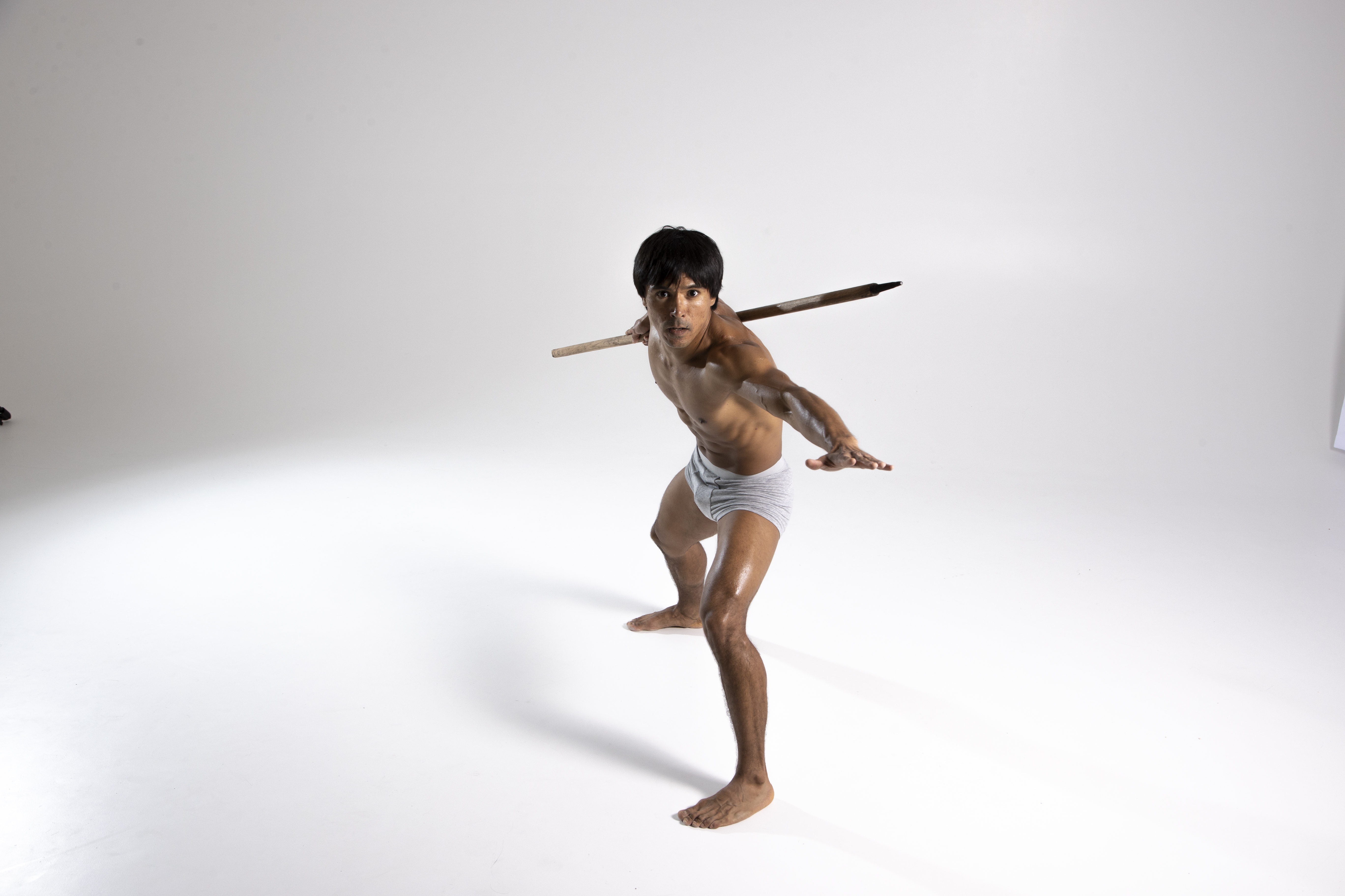a shirtless man is posed in a studio against a white background, holding a spear over his shoulders. he is wearing only a gray loincloth and stands barefoot in an action pose, suggestive of a defensive stance or martial arts. his athletic build and the traditional weapon create the impression of a warrior or fighter. the stark white backdrop keeps the focus on the figure and the spear.