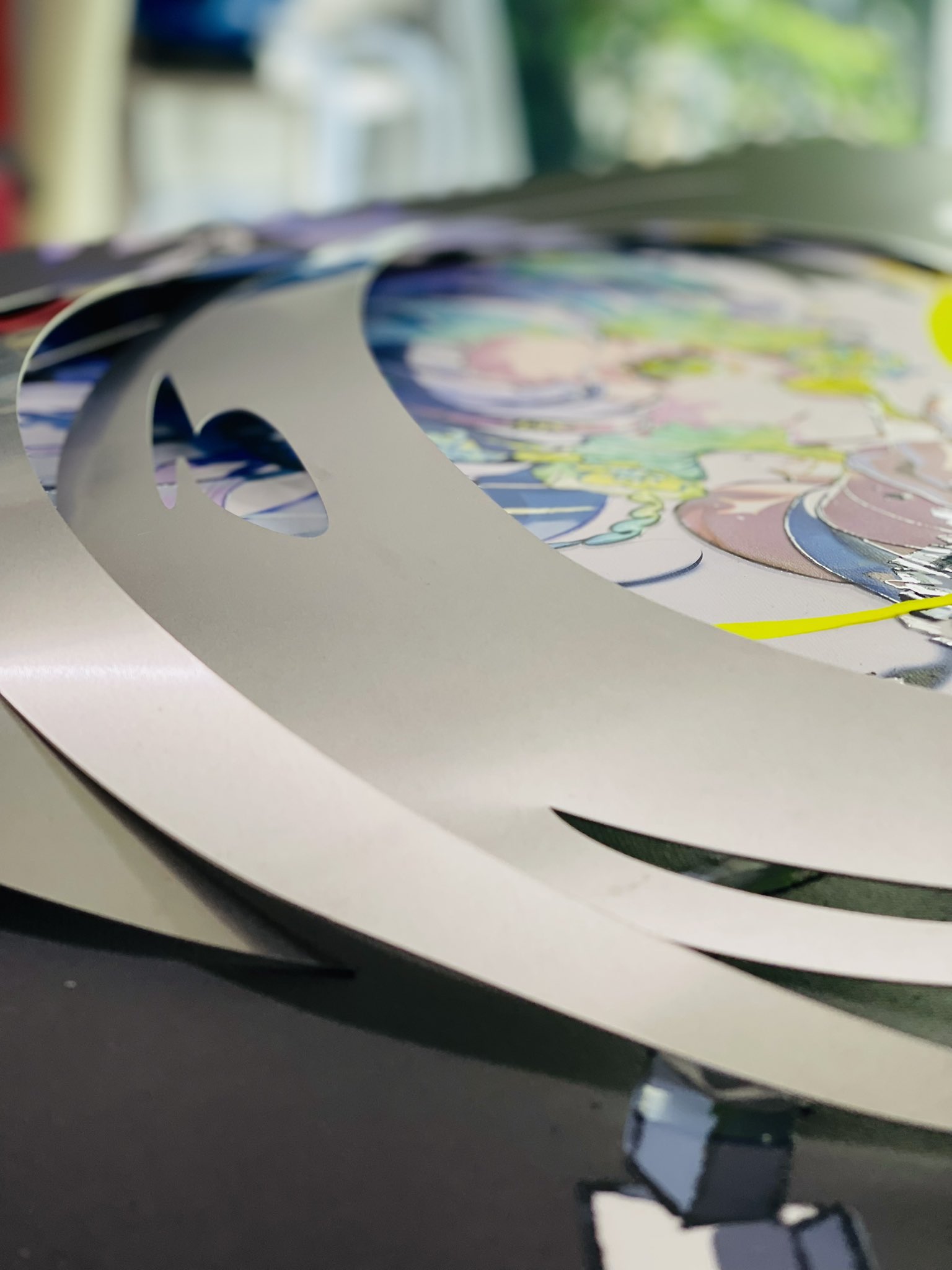 a closeup photo of a stack of silver vinyl decals featuring anime characters. the decals are cut into curved shapes and appear to be intended for use on a car. the background of the image is blurred, but suggests a workshop or studio setting.