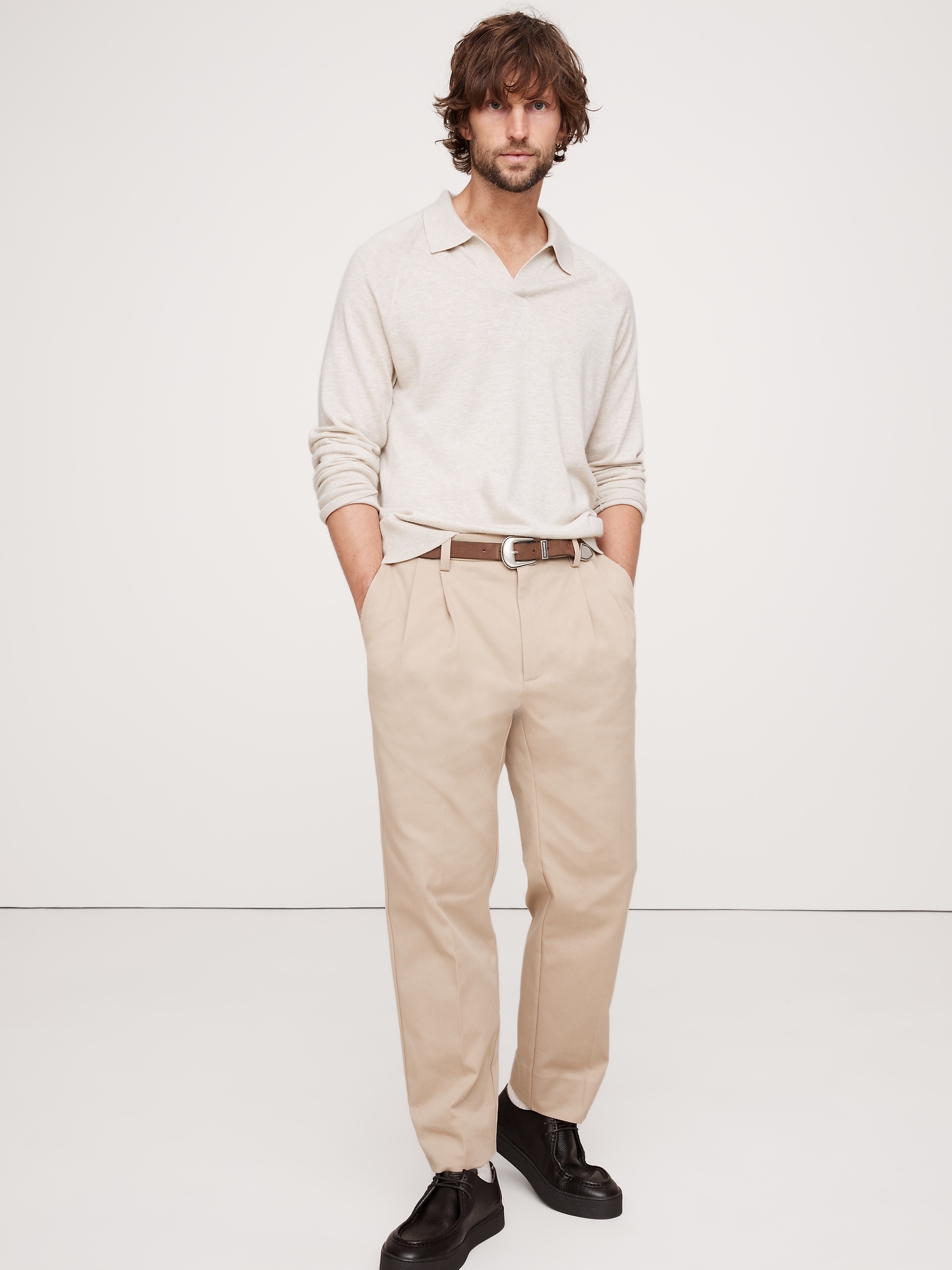 a fulllength studio shot of a man in a casual outfit. he's wearing beige pants with a brown belt, a lightcolored longsleeved polo shirt, and black shoes. he has mediumlength brown hair and is standing with his hands in his pockets against a white background.