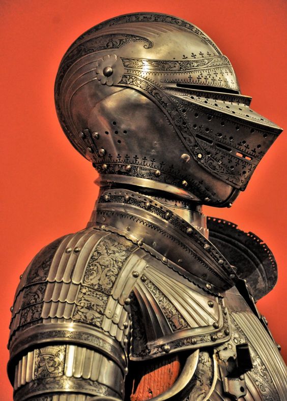 a closeup view of an ornate suit of medieval armor displayed against a vibrant orange background. the armor, crafted from polished steel, is intricately engraved with floral and geometric designs. the helmet, with its raised visor, is particularly striking. the image highlights the craftsmanship and artistry of historical armor.