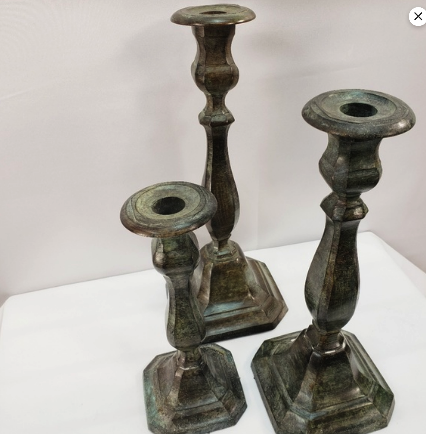 a set of three antique metal candlesticks, each with a different design and a greenbrown patina. the candlesticks are arranged on a white surface. the one in the back has a square base and a tall cylindrical stem with a circular top. the one on the left has a similar base, but its stem is shorter, with a round, flat top. the one on the right has a round base and a wide, flat stem with a square top. the image is taken from a slightly elevated angle, showing the candlesticks in a clear and welllit environment.