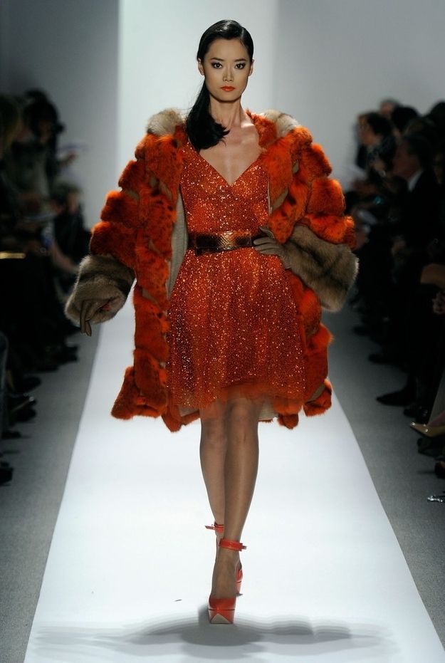 a woman in a red sequined dress and a fur coat walks down the runway. she has her hair pulled back and is wearing red shoes.  the runway is white and there is a crowd of people in the background.