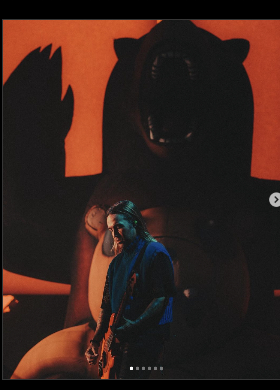 a man in a blue sweater and black pants plays a bass guitar on a stage lit by a spotlight. he is standing in front of a large inflatable bear, which is illuminated from behind with red light. the man is focused on his playing, and he is surrounded by darkness.