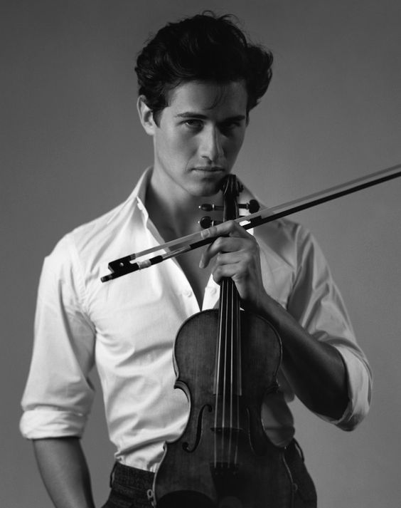 a black and white portrait of a young man in a white shirt, holding a violin. he is looking directly at the camera with a serious expression. the background is a plain, light gray. the lighting is soft and diffused, creating a sense of intimacy and drama.