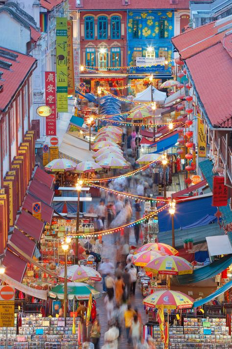 the image is an overhead, slightly angled shot of a bustling night market in singapore's chinatown. the street is lined with colorful shophouses, many adorned with chinese lanterns and signage. numerous market stalls, shaded by brightly colored umbrellas, fill the street, offering a variety of goods and street food. a crowd of people moves through the market, their forms slightly blurred due to motion. the overall scene is vibrant and energetic, showcasing the vibrant culture and atmosphere of a typical singaporean night market.  the lighting suggests it's dusk or early evening. the architecture of the buildings surrounding the market also contributes to the image's rich cultural context.