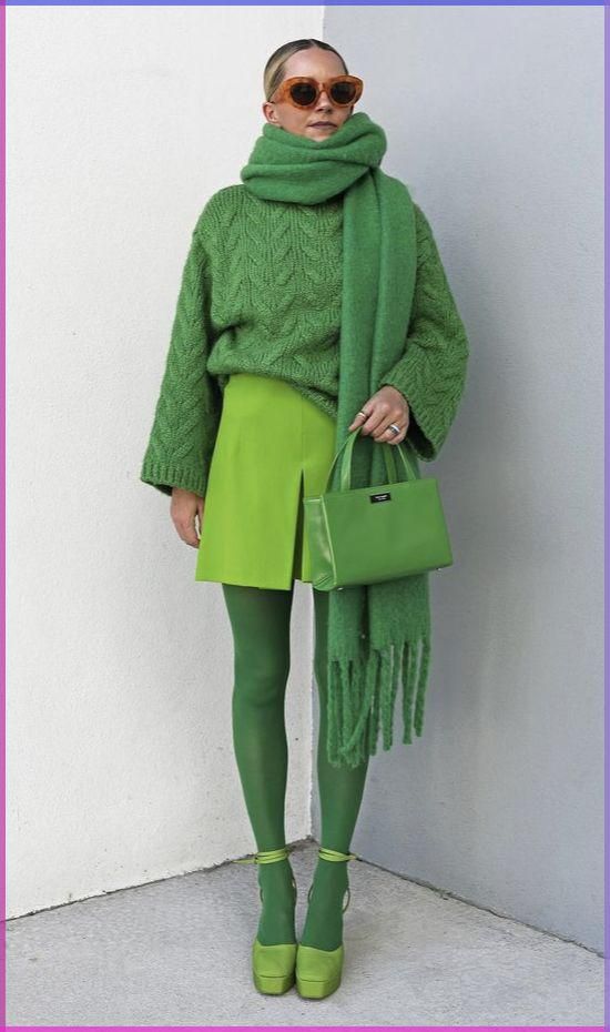 a woman in a green sweater, skirt, tights, and shoes poses with a green bag and scarf, looking at the camera. she is wearing sunglasses. the photo is taken in front of a plain white wall.