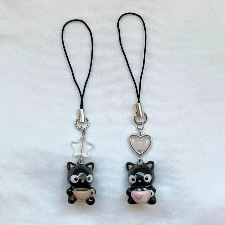 two black cat keychains with a black strap and metal attachment are displayed on a white fabric surface. the left keychain has a clear star charm, and the right keychain has a pink heart charm. each cat is holding a cup.