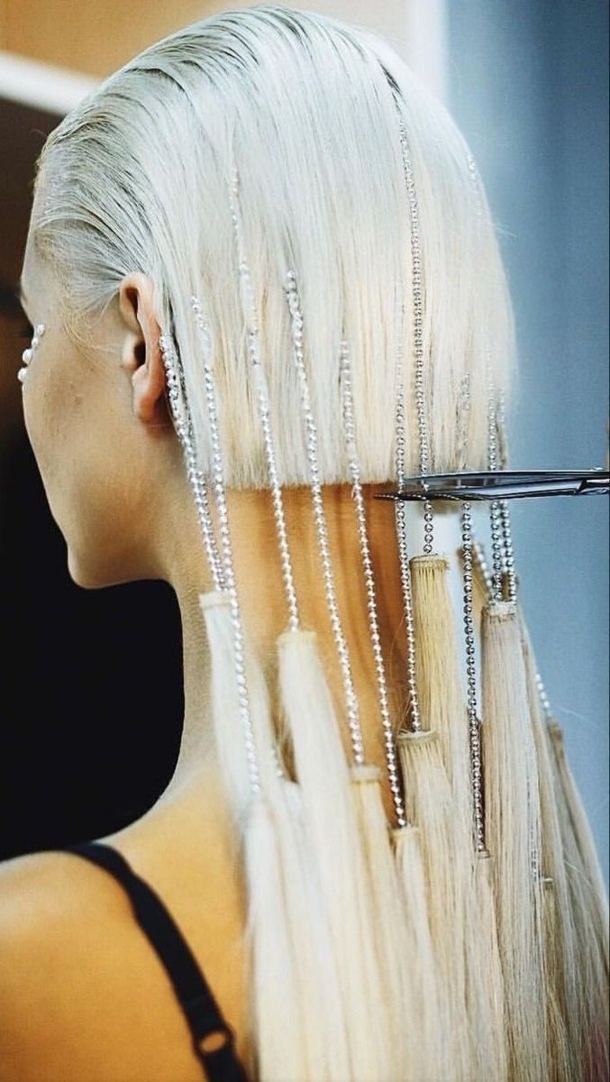 a blonde woman is getting her hair cut by a stylist. she is wearing a black top and has her long blonde hair tied up with a silver beaded hair accessory. the stylist is using a pair of scissors to cut her hair. the image is very glamorous and stylish.  the picture appears to be taken in a salon setting, with the woman's face partly obscured by the long strands of blonde hair, and the stylist's hand and arm are cropped out of the picture.