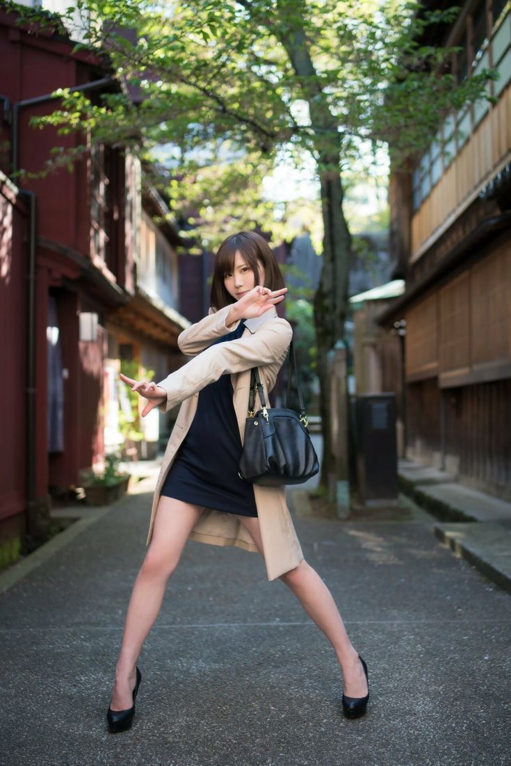 a young japanese woman in a beige coat and black dress poses stylishly on a city street. she is standing in a narrow alleyway with old buildings on either side. she is wearing a long beige coat, a black dress, and black high heels. she has long, black hair and is carrying a black handbag over her shoulder. she is looking directly at the camera with a confident expression. the photo is wellcomposed and highlights the woman's fashion sense and style.