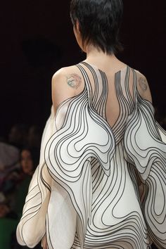 a fashion model is wearing an abstract, white dress with black lines. the dress has a flowing, organic design that resembles swirling, layered curves. the model has short black hair and is shown from the back, highlighting the intricate details of the dress's pattern and texture. the dress is a contemporary and avantgarde piece that showcases unique artistic expression.