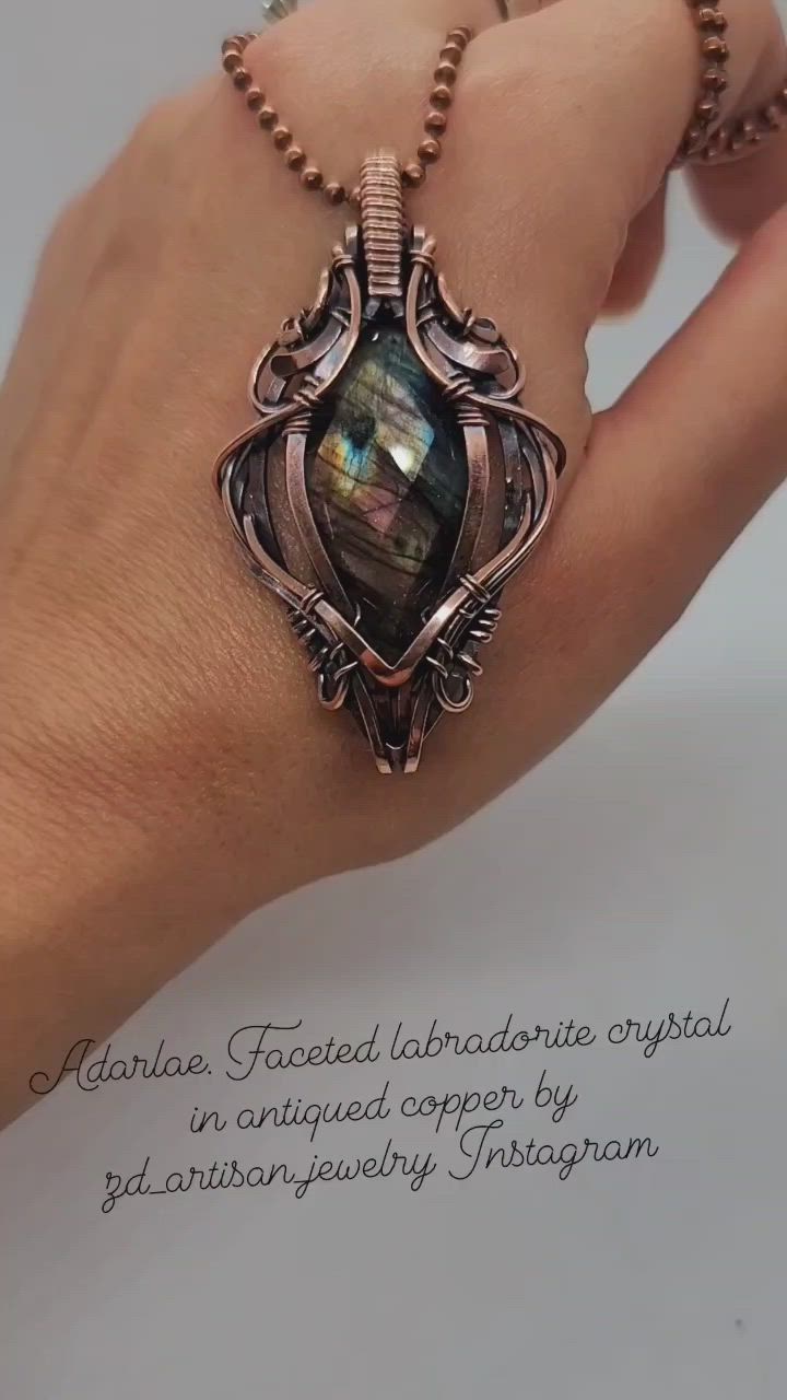 a closeup image shows a handmade wirewrapped pendant necklace featuring a faceted labradorite crystal set in antiqued copper. the pendant is being held in someone's hand, showcasing its size and detail. the chain is made of copper beads. text at the bottom indicates the pendant is made by zd_artisanjewelry on instagram.