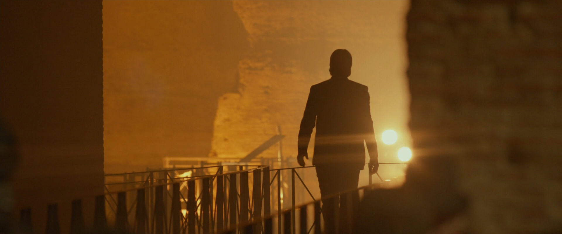 a man in a suit walks away from the camera, silhouetted against the smoky, warmly lit interior of a large space. he's on a metal walkway with a railing on his left, which is also silhouetted against the light. there's a hint of industrial equipment in the background. the overall mood is mysterious and suspenseful.