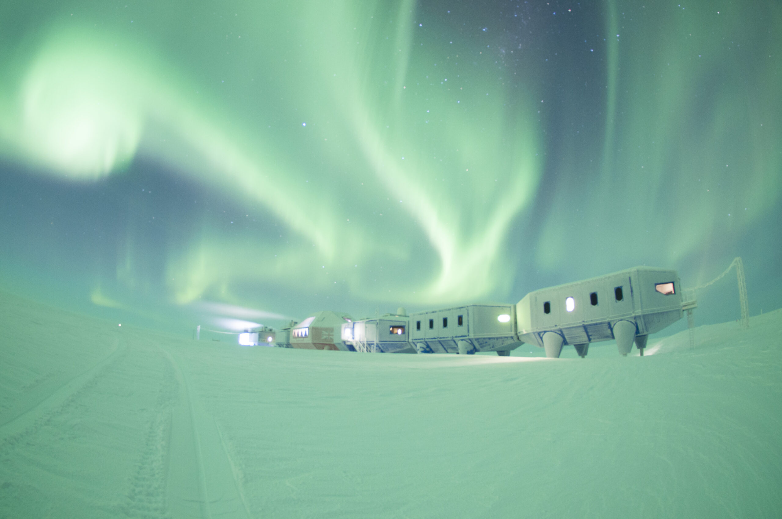 a row of white buildings sit under an amazing display of green aurora borealis in the night sky. the sky is filled with stars and the landscape is covered in snow and ice. the scene is peaceful and serene, with a sense of vastness and solitude.