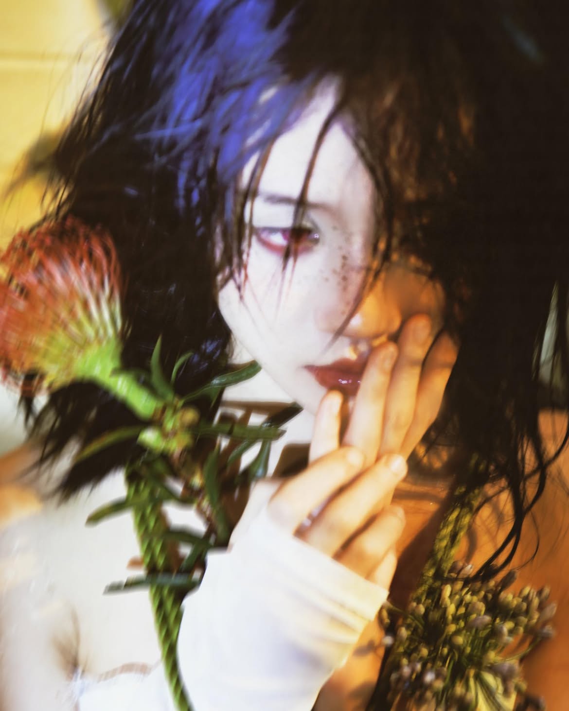 a blurred portrait of a woman with dark hair, fair skin, and red eyes. she has freckles and is wearing red lipstick. her hand is near her face, with her fingers touching her lips. there are flowers near her face and a piece of white fabric covering her shoulder. the image has a blurred and slightly surreal quality.